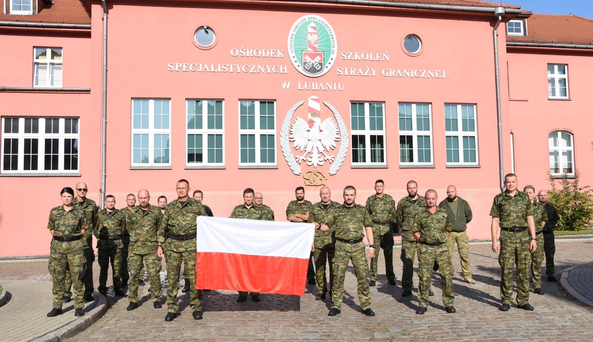Grupa funkcjonariuszy Straży Granicznej stojąca na tle budynku w mundurach polowych z flagą państwową pozująca do zdjęcia.