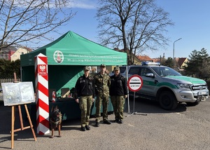 Funkcjonariusze SG przed stoiskiem promocyjnym Troje funkcjonariuszy SG stoją przed stoiskiem promocyjnym SG. Obok stoi samochód służbowy SG