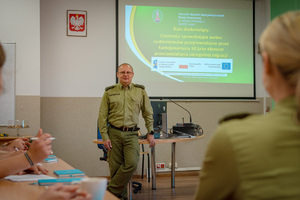 Kurs doskonalący pt. „Czynności sprawdzające wobec cudzoziemców przeprowadzane przez funkcjonariuszy SG jako element przeciwdziałania nielegalnej migracji” 02_09.05.2025_Funkcjonariuszka SG siedząca tyłem, w tle prowadzący szkole